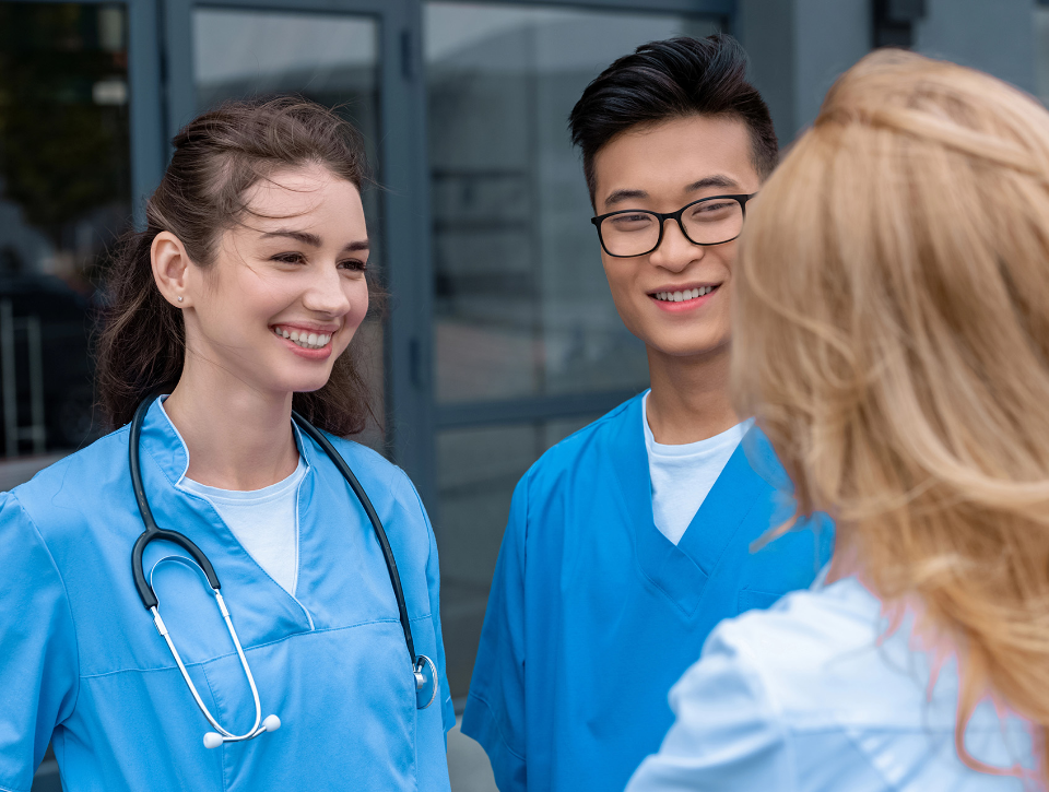 Deux étudiantes et un étudiant en sciences de la santé, en uniforme médical bleu, discutent en souriant à l’extérieur d’un bâtiment
